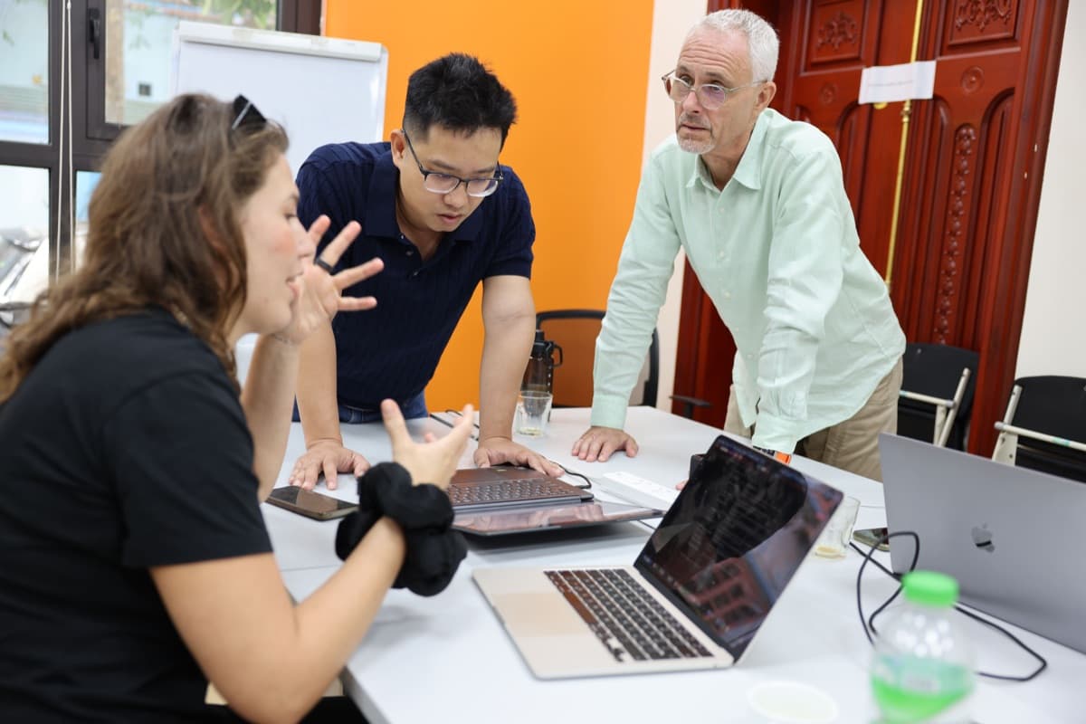Three people leaning into one problem across a table of laptops, mid-discussion